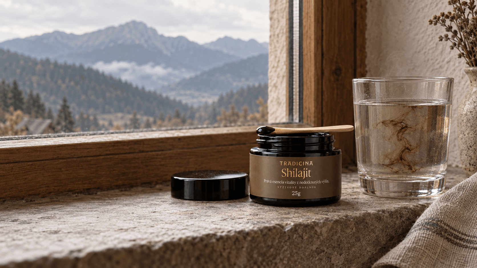 Otvorená čierna dóza Shilajit TRADICINA 25 g s drevenou lyžičkou na kamennom parapete, vedľa pohár teplej vody s rozpúšťajúcou sa živicou a výhľad na hory v opare