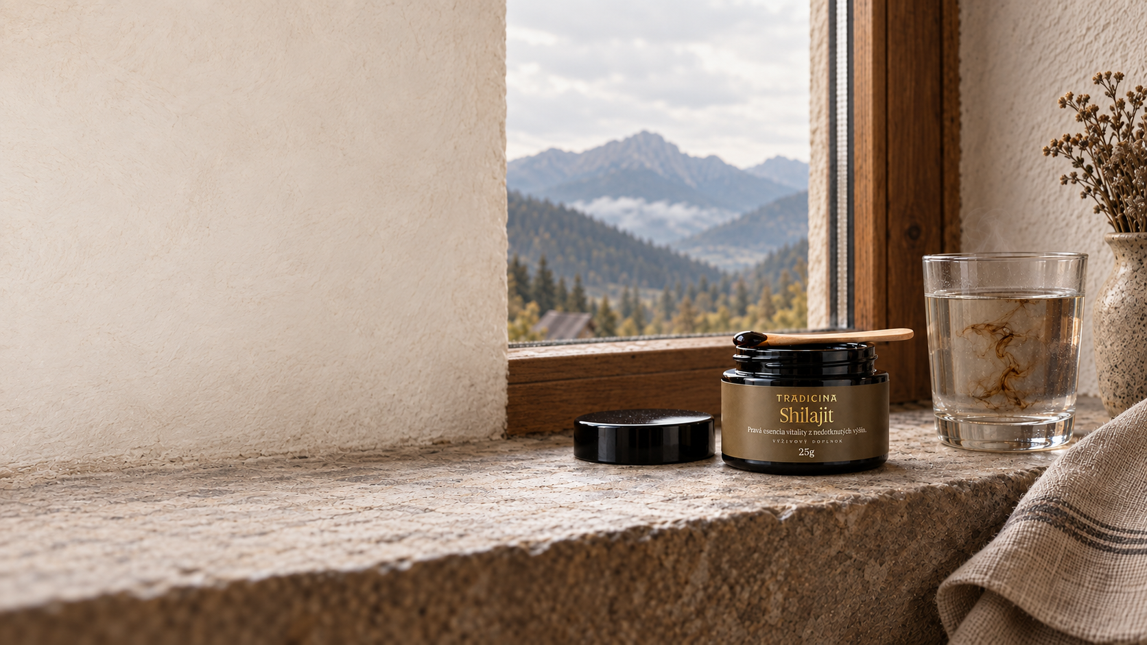 Otvorená čierna dóza Shilajit TRADICINA 25 g s drevenou lyžičkou na kamennom parapete, vedľa pohár teplej vody s rozpúšťajúcou sa živicou a výhľad na hory v opare